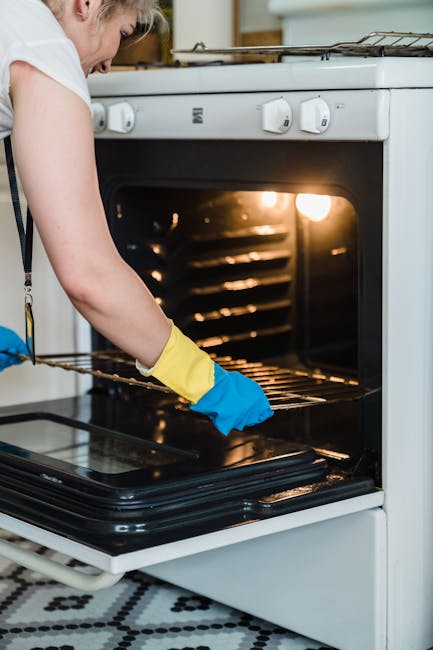 Close-up of a gloved hand cleaning the black ceramic surface of an oven with a blue cloth, removing soap suds and grease. The oven door is open, revealing the stainless steel interior racks and the ov